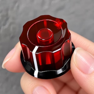 May include: A translucent red circular control knob with a scalloped edge and a flat top. The knob features a vertical white indicator line running down the side and is mounted on a solid black circular base with a smooth finish.