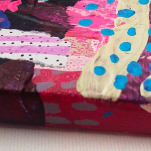 May include: Close-up of a colorful abstract painting with pink, blue, white, and brown paint. The painting has a textured surface and a variety of patterns, including stripes, dots, and swirls.