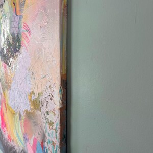 May include: Abstract painting on canvas with a textured surface. The painting features a variety of colors, including pink, yellow, blue, and green. The edges of the canvas are visible.