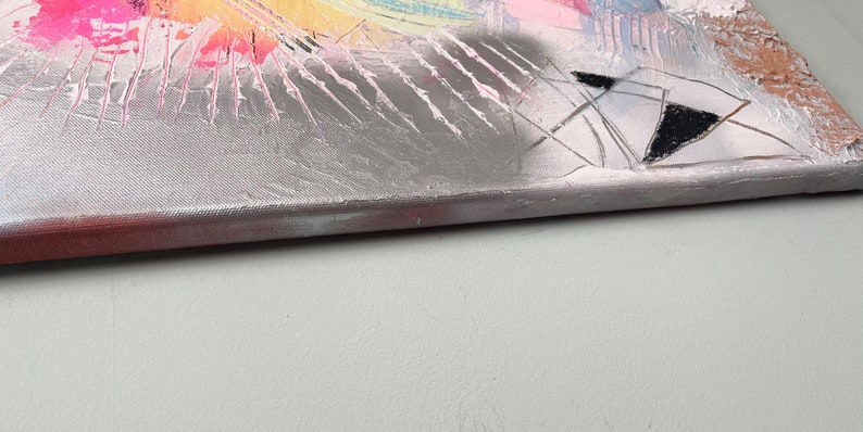 May include: Close-up of the edge of a silver canvas painting with abstract shapes and colors. The canvas is textured and has a pink, white, and black color scheme.