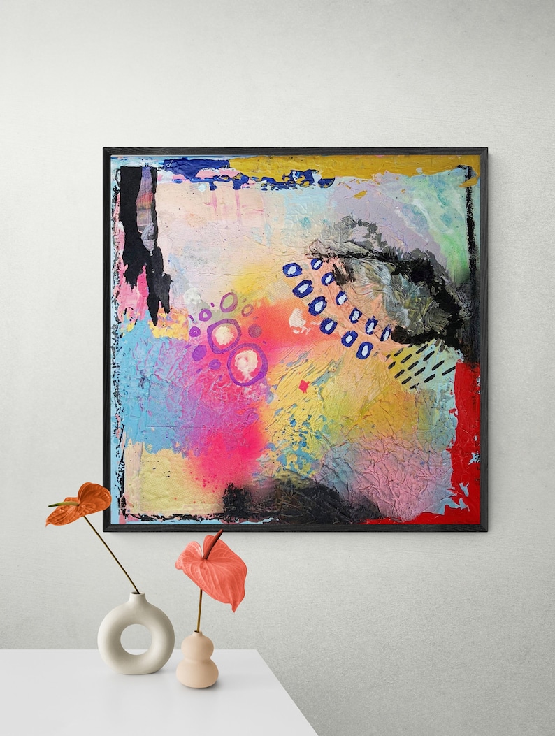 May include: Abstract painting with a black frame. The painting features a colorful background with pink, blue, yellow, and white paint splatters. There are also blue circles and black lines throughout the painting.