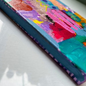 May include: A small, colorful abstract painting on canvas with a blue, green, pink, yellow, orange, and purple color scheme. The painting has a textured surface and is framed with a light blue border.