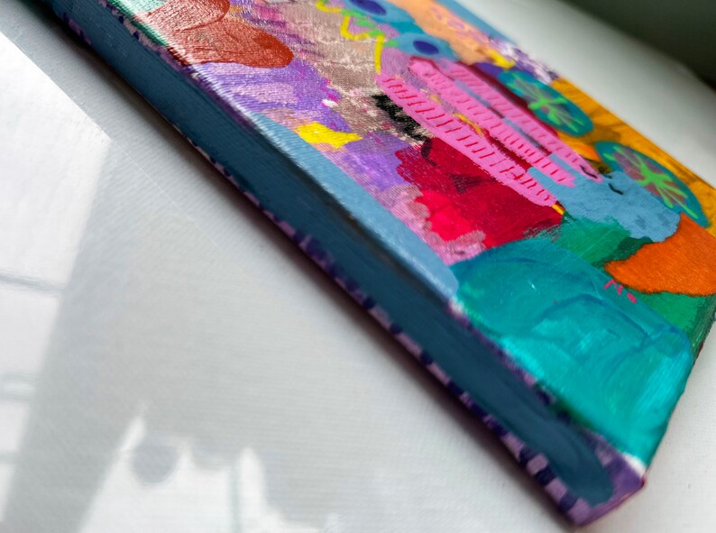 May include: A small, colorful abstract painting on canvas with a blue, green, pink, yellow, orange, and purple color scheme. The painting has a textured surface and is framed with a light blue border.