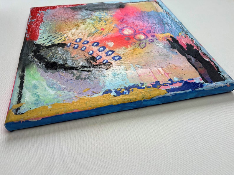 May include: Abstract painting on canvas with a textured surface. The painting features a mix of colors, including red, pink, blue, yellow, and black. The edges of the canvas are painted blue and gold.