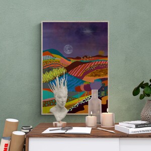 May include: A framed abstract painting of a colorful landscape with a moon and stars in the night sky. A white ceramic sculpture of a woman's head with a spiky crown is in front of the painting on a wooden table. There are two candles, a glass vase, and a magnifying glass on the table.