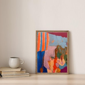 May include: A framed abstract painting featuring vertical orange and blue stripes, a large mauve shape, and patches of dark blue, green, and red. The artwork is displayed in a thin wooden frame, positioned next to a stack of books and a ceramic mug.