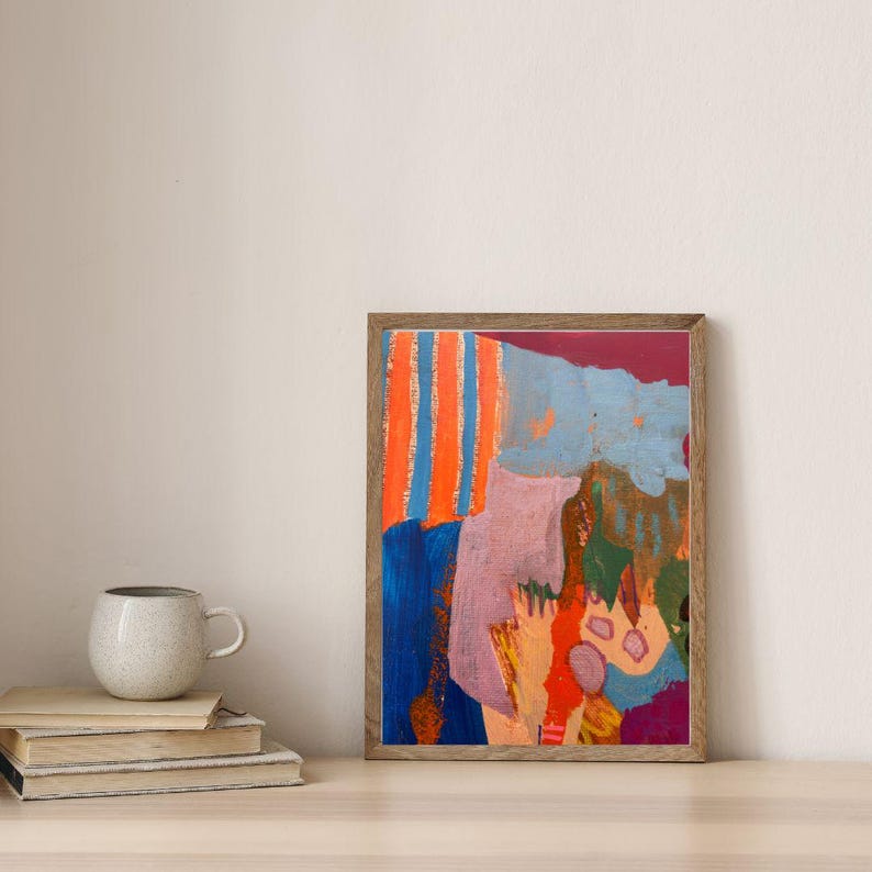 May include: A framed abstract painting featuring vertical orange and blue stripes, a large mauve shape, and patches of dark blue, green, and red. The artwork is displayed in a thin wooden frame, positioned next to a stack of books and a ceramic mug.