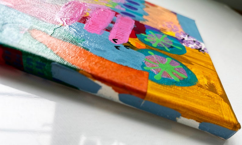 May include: A small, colorful abstract painting on canvas. The painting features a variety of shapes and colors, including pink, blue, green, orange, and yellow. The painting is framed with a blue border.