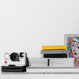 Op de afbeelding: Een witte instant camera met een rode knop en een stapel boeken en een ingelijst abstract schilderij met een kleurrijk gezicht op een witte achtergrond.