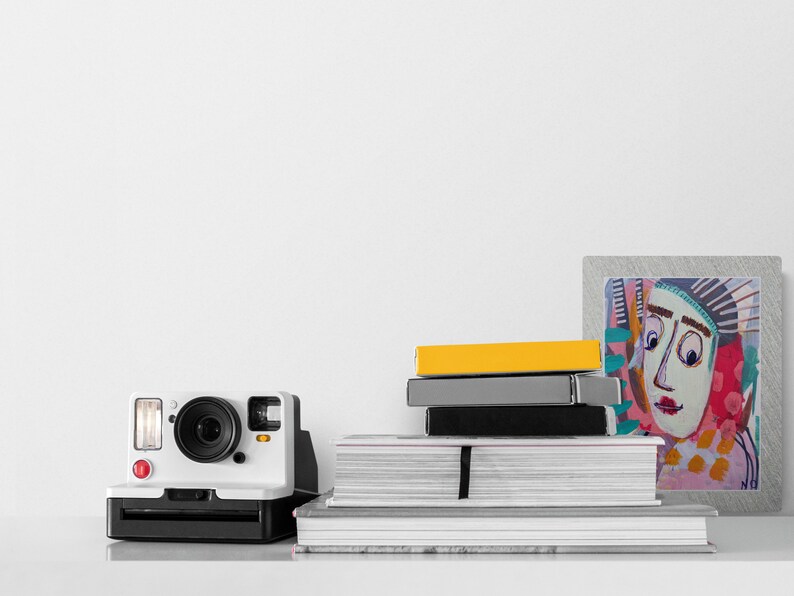 Op de afbeelding: Een witte instant camera met een rode knop en een stapel boeken en een ingelijst abstract schilderij met een kleurrijk gezicht op een witte achtergrond.