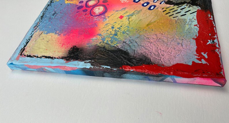 May include: Abstract painting on canvas with a textured surface. The painting features a variety of colors, including pink, blue, yellow, red, and black. The edges of the canvas are painted in a light blue color.
