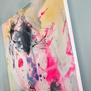 Puede incluir: Pintura abstracta sobre lienzo con salpicaduras de pintura rosa, amarilla, azul y negra. La pintura tiene una superficie texturizada y un fondo blanco.
