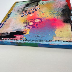 May include: Abstract painting on canvas with a colorful, textured background. The painting features a variety of colors, including pink, blue, yellow, and green. There are also black and white dots and circles throughout the painting.
