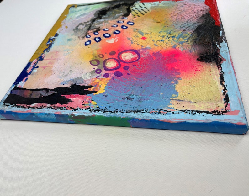 May include: Abstract painting on canvas with a colorful, textured background. The painting features a variety of colors, including pink, blue, yellow, and green. There are also black and white dots and circles throughout the painting.