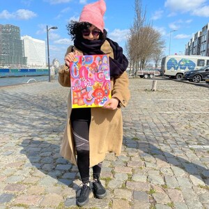 Puede incluir: Una persona con un gorro rosa, una bufanda negra y un abrigo marr&oacute;n sostiene una pintura abstracta colorida. La pintura est&aacute; sobre un fondo blanco y presenta una variedad de formas y colores, incluyendo rosa, amarillo, azul y verde.