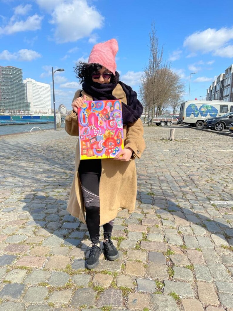 Puede incluir: Una persona con un gorro rosa, una bufanda negra y un abrigo marr&oacute;n sostiene una pintura abstracta colorida. La pintura est&aacute; sobre un fondo blanco y presenta una variedad de formas y colores, incluyendo rosa, amarillo, azul y verde.
