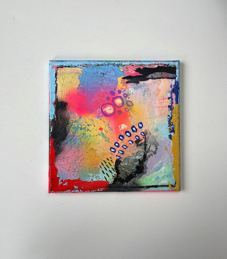 May include: Abstract painting on canvas with a textured surface. The painting features a mix of colors, including pink, blue, yellow, and black. There are also some white circles and blue dots on the canvas.