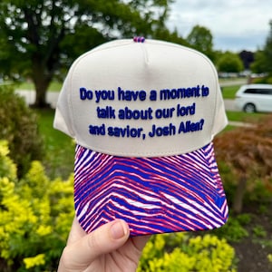 May include: A cream-coloured baseball cap featuring the embroidered blue text "Do you have a moment to talk about our lord and saviour, Josh Allen?" on the front panel, paired with a vibrant red and blue zebra-striped pattern on the brim.