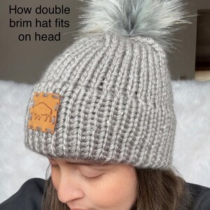 May include: A grey knitted beanie with a faux fur pom pom on top. The beanie has a brown leather patch with a buffalo silhouette on the side. The beanie is being worn by a person.