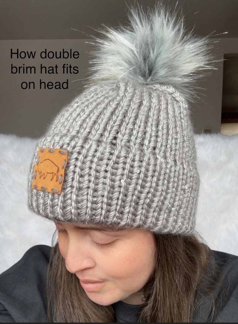 May include: A grey knitted beanie with a faux fur pom pom on top. The beanie has a brown leather patch with a buffalo silhouette on the side. The beanie is being worn by a person.