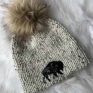 May include: A knitted beanie in a speckled light gray color with a large, fluffy, light brown pom-pom. The beanie features a black buffalo silhouette on the front. The pom-pom has gold metallic strands woven throughout.