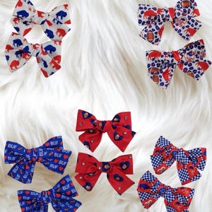 May include: A collection of fabric hair bows in various patterns and colors. Some bows feature buffalo and football designs in red, white, and blue. Other bows have the word "BUF" and heart shapes. The bows are arranged on a white, fluffy surface.