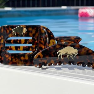 May include: Two hair clips with a brown and black tortoiseshell pattern. Each clip features a gold-toned metal buffalo emblem centered on the surface. The rectangular clip has three horizontal cutouts, while the curved clip has a claw-style design.