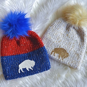 May include: Two knitted winter hats with pom poms. One hat is red and blue with a white buffalo silhouette. The other hat is white with gold glitter and a gold buffalo silhouette.