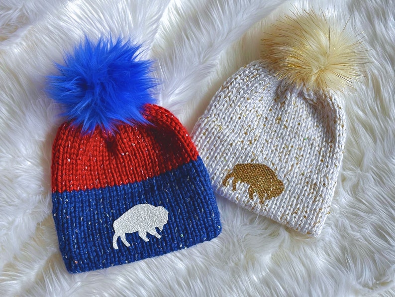 May include: Two knitted winter hats with pom poms. One hat is red and blue with a white buffalo silhouette. The other hat is white with gold glitter and a gold buffalo silhouette.