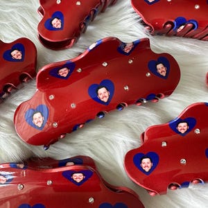 May include: Several red hair claw clips with a glossy finish, featuring blue heart-shaped images of a man's face and clear rhinestones. The clips have a curved design and are displayed on a white, fluffy surface.