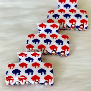 May include: Three white plastic hair clips with a repeating pattern of red and blue buffalo silhouettes.