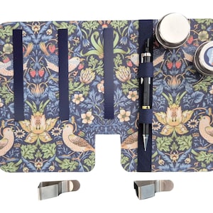May include: A rectangular folding organizer featuring a dark blue floral and bird pattern with strawberries. It includes four vertical elastic bands, a pen holder with a black pen, two circular metal containers, and two separate metal clips.