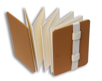 May include: An accordion-style book with solid tan covers and cream-colored pages. The book features a white elastic band closure that wraps around the front cover to keep the pages securely folded together in a concertina arrangement.