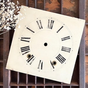 May include: A square, off-white clock face with black Roman numerals and hands. The clock face has two small holes and a central hole. The clock face is slightly angled, revealing a glimpse of a wooden background.