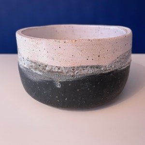 Könnte beinhalten: Eine weiße und schwarze Keramikschüssel mit gesprenkelter Textur. Die Schüssel hat einen breiten Rand und einen abgerundeten Boden.