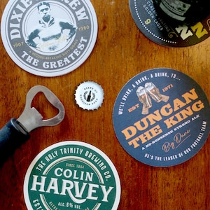 Puede incluir: Cuatro posavasos de cerveza con diferentes diseños. Los posavasos son redondos y presentan diferentes logotipos y textos. Los posavasos están sobre una superficie de madera.