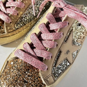 May include: Close-up of a pair of gold and silver glitter sneakers with pink laces. The sneakers have a star design on the side.