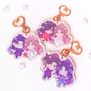 May include: Three sets of anime character keychains. Each keychain features two characters, one male and one female, with a heart-shaped gold metal ring. The characters are drawn in a chibi style and have colorful clothing and hair.
