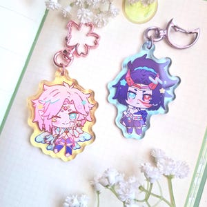 Puede incluir: Dos llaveros de personajes de estilo anime. Un llavero presenta un personaje de pelo rosa con un contorno amarillo y un cierre en forma de flor. El otro llavero tiene un personaje de pelo morado con un contorno azul y un cierre plateado.
