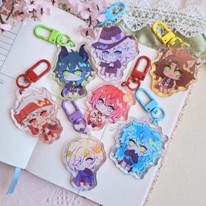Peut inclure: Une collection de porte-clés de personnages de style anime avec des designs colorés. Chaque porte-clés présente un personnage unique avec des cheveux et des tenues vibrantes, attaché à un fermoir en métal. Les porte-clés sont exposés sur un carnet ouvert.