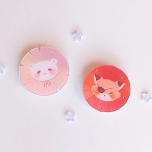 May include: Two round pins with a white background and a colorful design. The first pin features a white bear wearing a pink hat with a pink and orange gradient background. The second pin features a red fox wearing an eye patch with a red and orange gradient background.