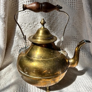 May include: A vintage brass teapot with a wooden handle and a curved spout. The teapot has a round body, a lid with a decorative finial, and small feet. The brass has a warm, golden color, and the handle is a dark brown.