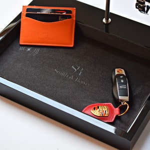 May include: Black valet tray with a gray felt lining. The tray has a wireless charging pad, a slot for keys, and a leather key fob with a Porsche logo. The tray is labeled "Smith & Jones".