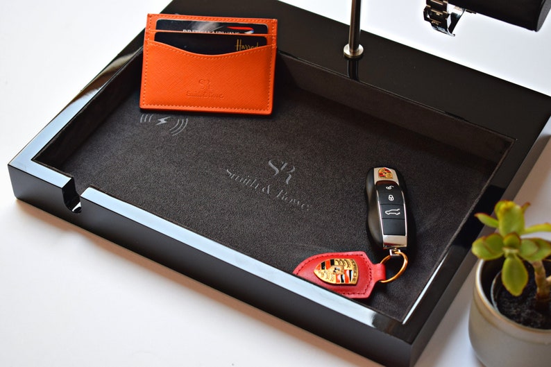 May include: Black valet tray with a gray felt lining. The tray has a wireless charging pad, a slot for keys, and a leather key fob with a Porsche logo. The tray is labeled "Smith & Jones".