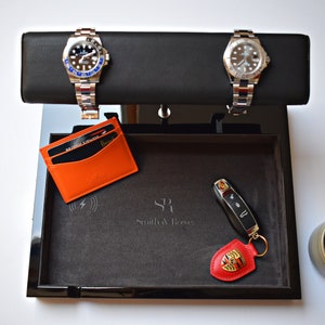 May include: A black valet tray with a grey felt lining. The tray contains two silver watches, a black key fob with a red Porsche logo, and a black and orange leather card holder. The tray also has a wireless charging pad and the text "SR Smith & Royce" in grey.