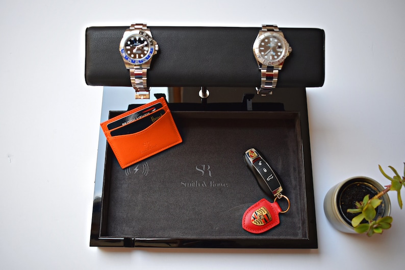 May include: A black valet tray with a gray felt lining. The tray contains two silver watches, a black key fob with a red Porsche logo, and a black and orange leather card holder. The tray also has a wireless charging pad and the text "SR Smith & Royce" in gray.