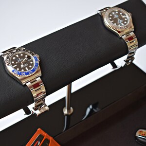May include: Two silver wristwatches with black faces and blue bezels are displayed on a black leather watch winder. The watch winder is attached to a black tray with a car key and a small orange wallet.