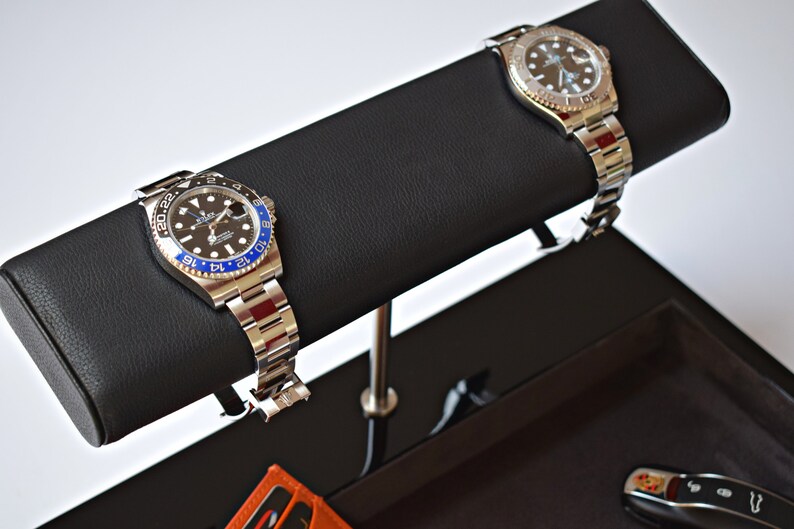 May include: Two silver wristwatches with black faces and blue bezels are displayed on a black leather watch winder. The watch winder is attached to a black tray with a car key and a small orange wallet.