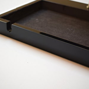 May include: A black rectangular tray with a black felt lining and a small slot on the side. The tray is made of wood and has a glossy finish.