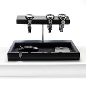 May include: A black valet tray with a grey felt lining. The tray has a built-in watch stand with three slots for watches. There is a key and a smartphone on the tray.